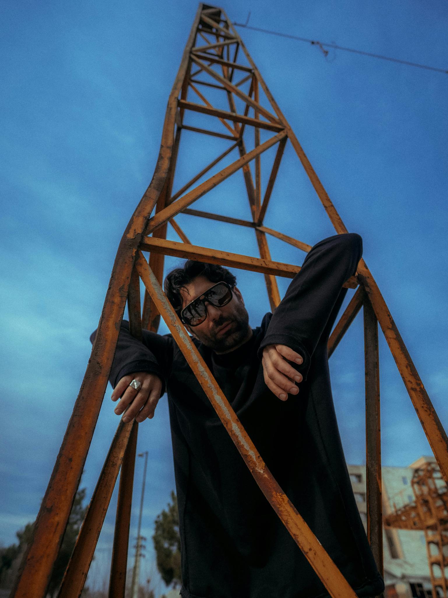 Man in sunglasses poses creatively by a metal framework against vibrant sky.